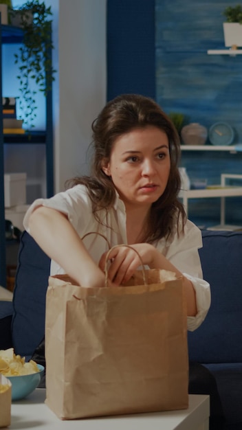 Adulto de raza caucásica con comida para llevar tomando comida de la bolsa a partir de la televisión sentado en el sofá en casa. Mujer con paquete de entrega en la mesa viendo la televisión y comiendo comida rápida en el sofá