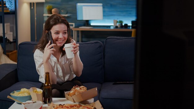 Adulto de raza caucásica disfrutando de una llamada telefónica con un amigo en la sala de estar mientras bebe café. Mujer hablando por teléfono inteligente, preparándose para comer comida de entrega de comida rápida en casa después del trabajo para relajarse