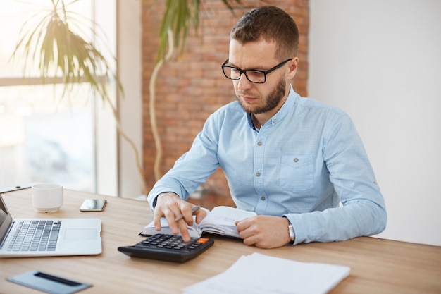 Adulto serio barbudo caucásico gerente de finanzas en gafas y camisa azul sentado en la cómoda oficina ligera