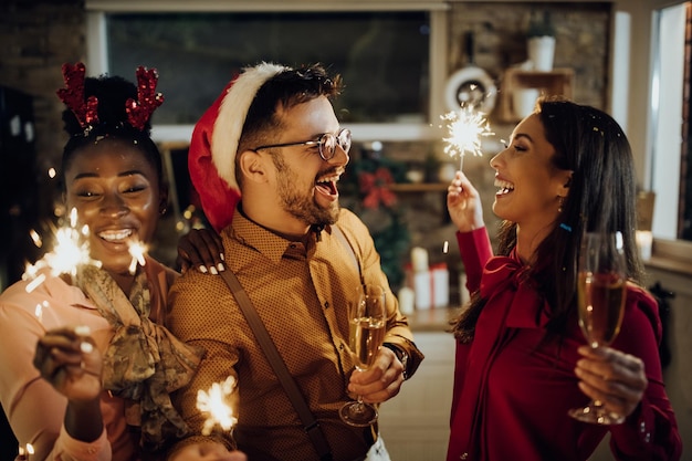 Adultos jóvenes alegres bebiendo champán y usando bengalas en la fiesta de Año Nuevo en casa