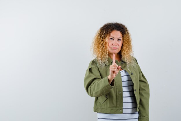 Advertencia de mujer bastante rubia con el dedo índice en chaqueta verde y mirando seria, vista frontal.