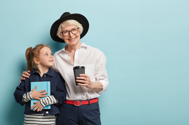 Afectuosa abuela de pelo gris con sombrero abraza a la pequeña niña, ama a su nieta, bebe café para llevar. Niño curioso con bolsillo, escucha consejos de anciana sabia.