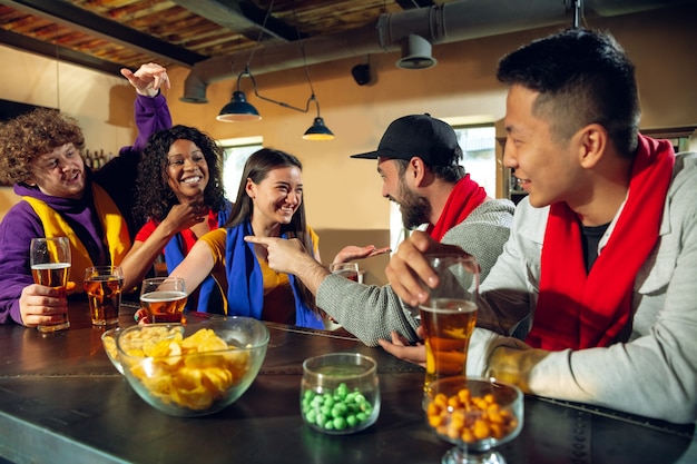 Aficionados al deporte animando en el bar, pub y bebiendo cerveza mientras se lleva a cabo el campeonato, la competencia. grupo multiétnico de amigos emocionado viendo la traducción. las emociones humanas, la expresión, el concepto de apoyo.