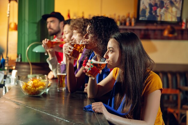 Aficionados al deporte animando en el bar, pub y bebiendo cerveza mientras se lleva a cabo el campeonato, la competencia. Grupo multiétnico de amigos.