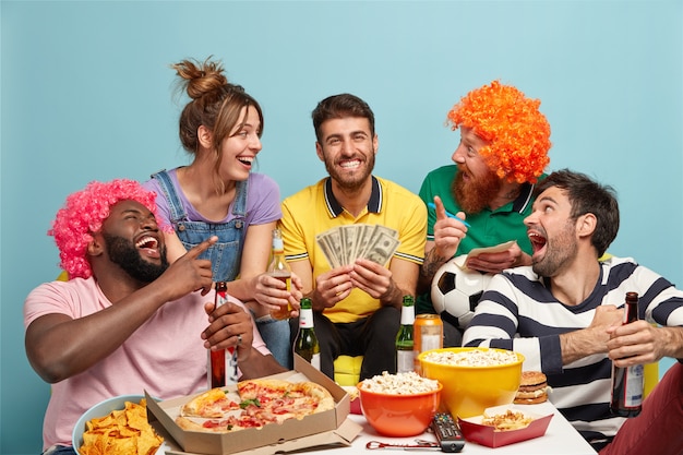 Aficionados al fútbol, concepto de felicidad y diversión. Amigo lleno de alegría contento de tener éxito en la apuesta de fútbol, ganar una suma global de dinero, guardar dólares, comer bocadillos sabrosos, sentarse a la mesa, reír a carcajadas, aislado en azul