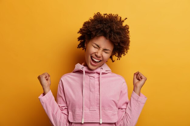 Afortunada y emocionada mujer bonita aprieta los puños, grita hurra, celebra las buenas noticias, inclina la cabeza, se viste informalmente, usa una sudadera con capucha rosa, disfruta de un dulce éxito, siente el sabor de la victoria, usa una sudadera de terciopelo