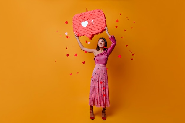 Afortunada y encantadora mujer de 23 años sostiene un cartel en forma de me gusta de Instagram y posa en pleno crecimiento en una pared naranja con confeti