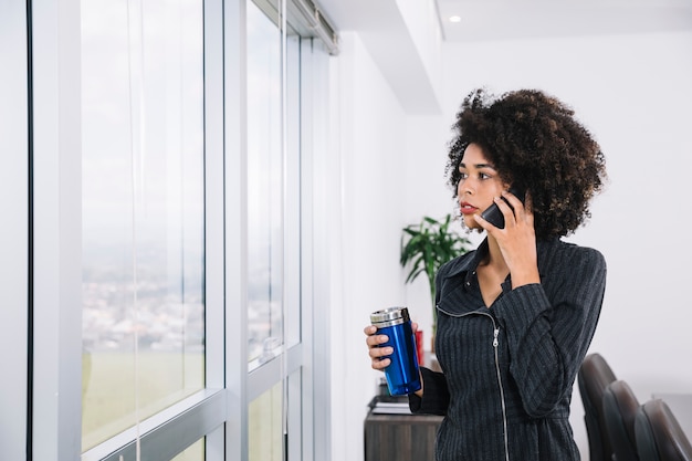 Afroamericana joven con termo hablando en teléfono inteligente cerca de la ventana