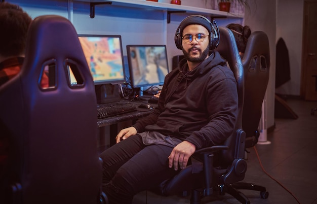 Foto gratuita un afroamericano barbudo con capucha y gorra sentado en una silla de jugador y mirando una cámara en un club de juegos o en un cibercafé.