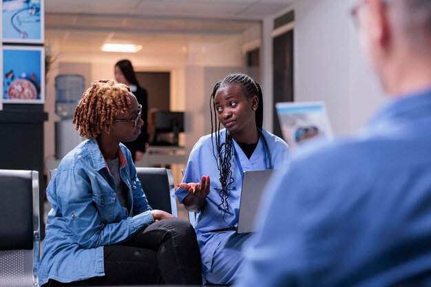 Afroamericanos haciendo consultas con diagnóstico de enfermedades en una computadora portátil, sentados en el vestíbulo de la sala de espera. Enfermera y paciente hablando sobre tratamiento y recuperación, apoyo sanitario en la clínica.