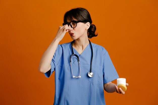 agarró la nariz sosteniendo la lata de píldora joven doctora vistiendo uniforme fith estetoscopio aislado sobre fondo naranja