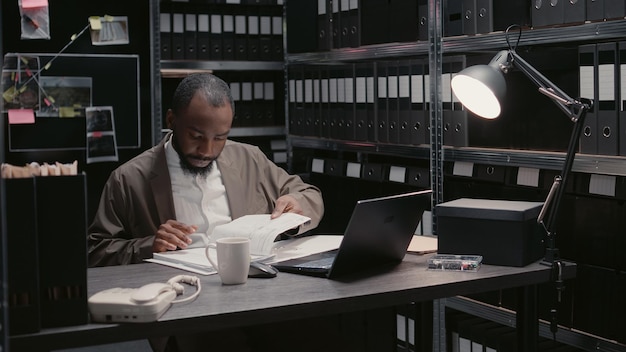 Agente afroamericano leyendo archivos sospechosos en la sala de archivo, tratando de resolver el crimen y atrapar al sospechoso. Inspector privado trabajando con mapa de detectives y pruebas forenses. Disparo de mano.