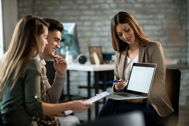 Agente inmobiliario sonriente usando una computadora mientras se reúne con una pareja joven y hace planes de inversión