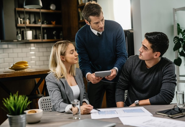 Agente inmobiliario trasero y pareja joven revisando planos y usando tableta digital en una reunión