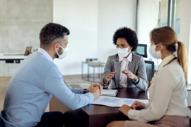 Agente de seguros afroamericana con mascarilla hablando con sus clientes en una reunión en la oficina