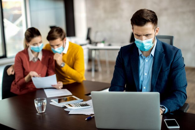 Agente de seguros con mascarilla trabajando en una laptop mientras sus clientes leen papeleo en segundo plano