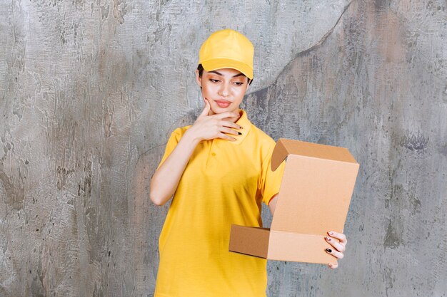 Foto gratuita agente de servicio femenino en uniforme amarillo sosteniendo una caja de cartón abierta y parece confundida y pensativa