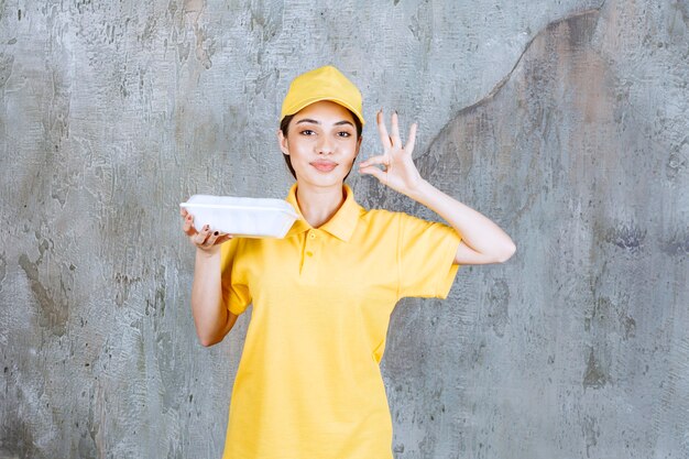 Agente de servicio femenino en uniforme amarillo sosteniendo una caja de comida para llevar de plástico y mostrando un signo de mano positivo.