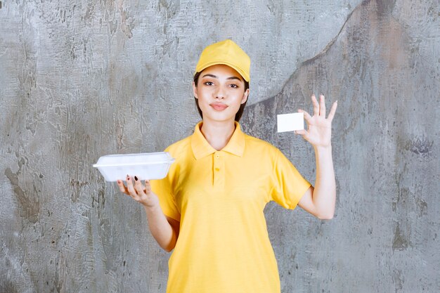 Foto gratuita agente de servicio femenino en uniforme amarillo sosteniendo una caja de comida para llevar de plástico y presentando su tarjeta de visita