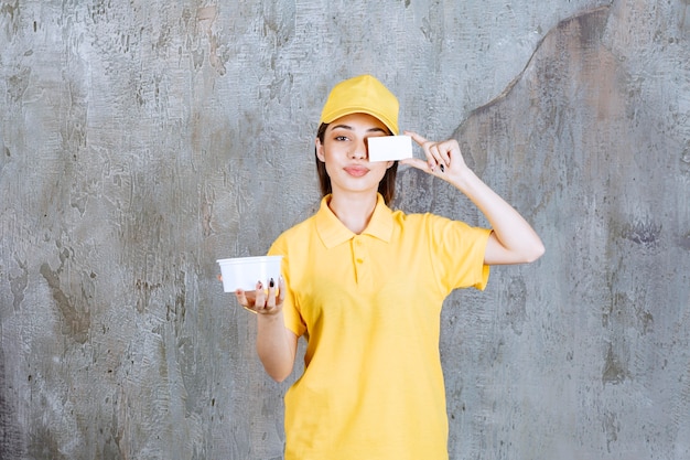 Agente de servicio femenino en uniforme amarillo sosteniendo un recipiente de plástico para llevar y presentando su tarjeta de visita.