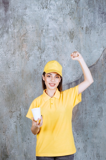 Agente de servicio femenino en uniforme amarillo sosteniendo un vaso de plástico y mostrando un signo de mano positivo.