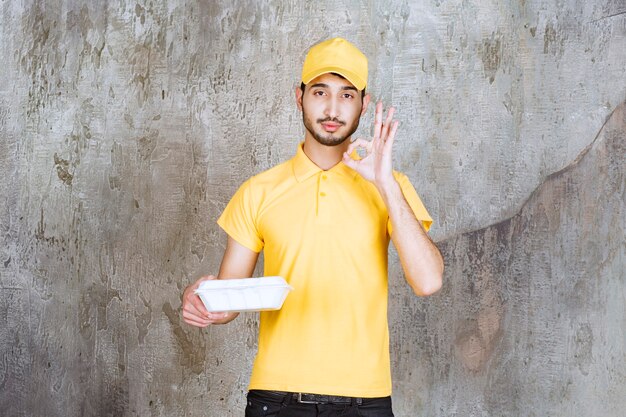 Agente de servicio masculino en uniforme amarillo sosteniendo una caja de comida para llevar blanca y mostrando un signo de disfrute.