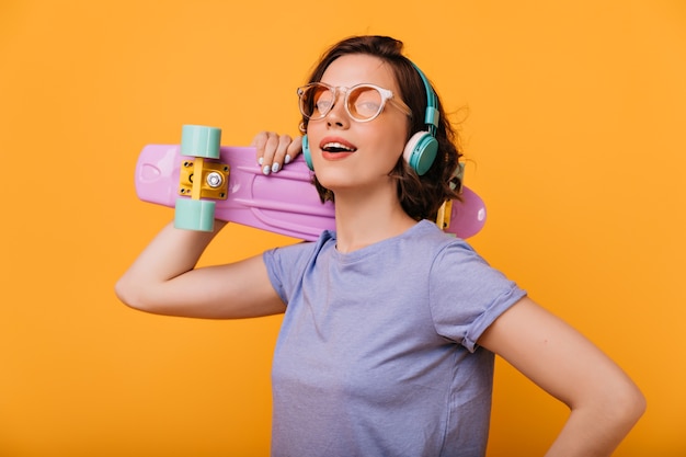 Agraciada chica blanca con patineta soñadora mirando a otro lado. Retrato de interior de hermosa mujer de pelo corto en auriculares azules.