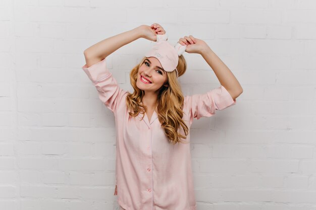 Agraciada chica con cabello ondulado posando en camisón de seda rosa. Mujer joven positiva en antifaz bailando en la mañana.