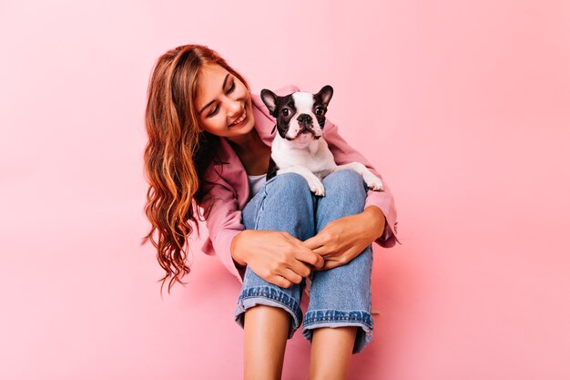 Agraciada chica de pelo largo mirando perro con amor. Señora alegre posando con bulldog francés de rodillas.