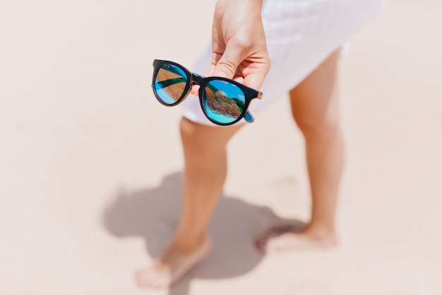 Agraciada dama descalza posando con gafas de sol. Retrato al aire libre de mujer con piernas bronceadas con gafas brillantes en primer plano.