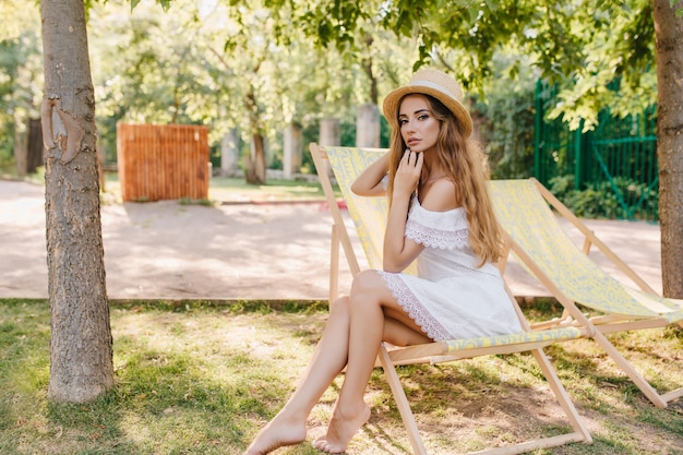 Agraciada dama descalza con sombrero de paja sentada en un sillón con expresión pensativa. Retrato al aire libre de una niña bonita de pelo largo con vestido blanco escalofriante en una silla en el parque.