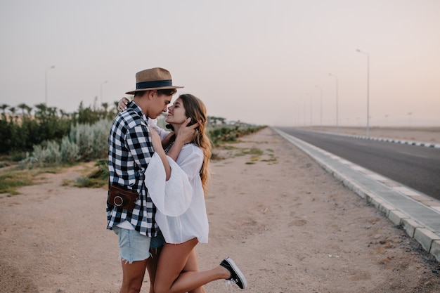 Agraciada mujer bonita en túnica blanca de pie sobre una pierna y abrazando suavemente a su novio cerca de la carretera. Hombre joven en pantalones cortos de mezclilla y sombrero de moda mirando a los ojos de su novia en cita al aire libre