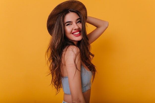 Agraciada mujer bronceada con elegante sombrero marrón de pie en pose de confianza y sonriendo