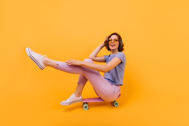 Agraciada mujer caucásica joven sentada en longboard rosa. Chica inspirada con cabello ondulado escuchando música favorita en elegantes auriculares.