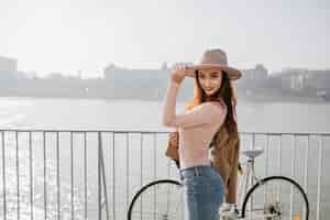 Foto gratuita agraciada mujer delgada en jeans tocando su sombrero, de pie en el puente de la ciudad