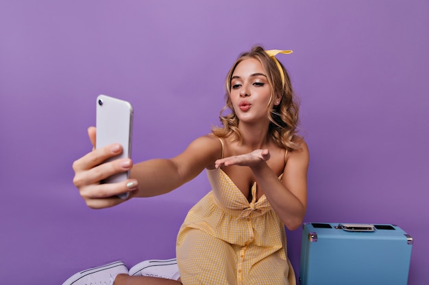 Agraciada mujer europea haciendo selfie antes de vacaciones. Retrato interior de hermosa niña caucásica en vestido amarillo sentada cerca de su maleta.