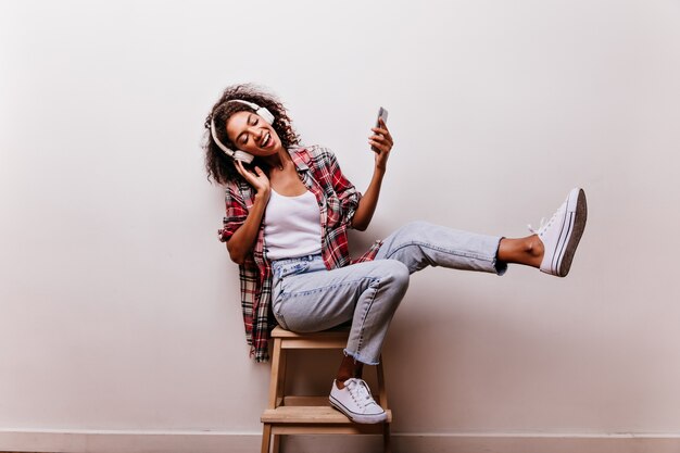 Agraciada mujer joven en jeans azul escuchando música en blanco. Filmación en interiores de una niña africana sin preocupaciones en auriculares sonriendo con los ojos cerrados.