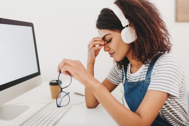 Agraciada mujer negra en auriculares con dolor de cabeza sentado al lado de la computadora y sosteniendo gafas en la mano