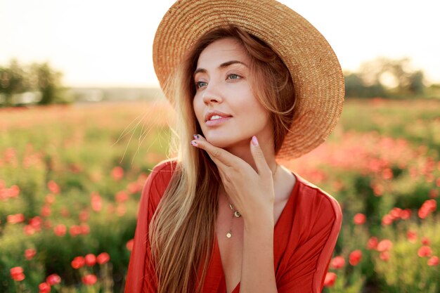 Agraciada mujer de pelo largo mirando al horizonte, disfrutando de la libertad. Chica seductora posando en campo de amapolas. Luz cálida del atardecer.