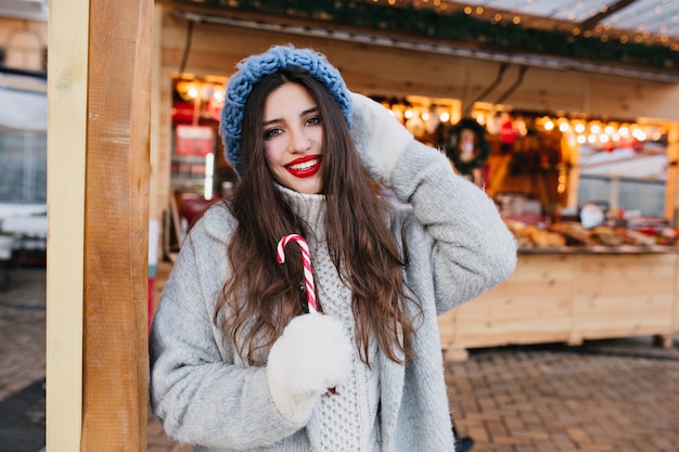 Agraciada mujer de pelo negro con bastón de caramelo sonriendo. Retrato al aire libre de la impresionante chica de moda en guantes blancos divirtiéndose en el mercado navideño.