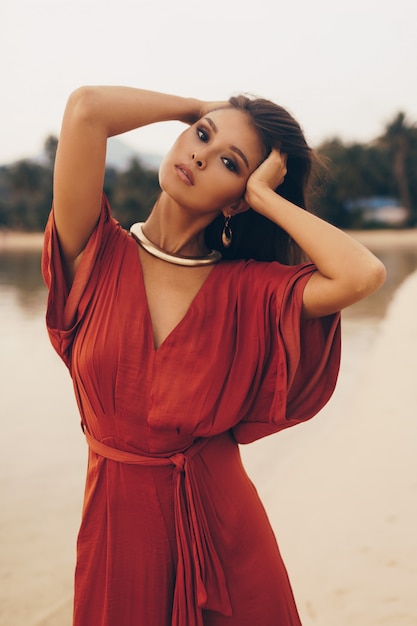 Agraciada mujer posando en la playa, sentada en la arena en vestido rojo