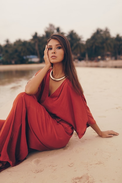Agraciada mujer posando en la playa, sentada en la arena en vestido rojo