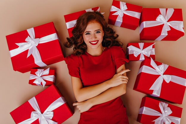 Agraciada mujer rizada de buen humor posando en el suelo con regalos. Modelo femenino atractivo que disfruta de la fiesta de Navidad.