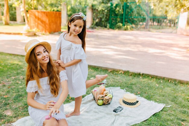 Agraciada mujer vestida de blanco sentada sobre una manta en el parque, mientras su linda hija baila a sus espaldas. Retrato al aire libre de dos hermanas alegres divirtiéndose después de un picnic.