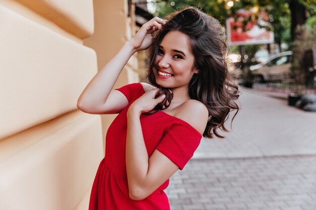 Agraciada niña feliz bailando en la calle. Debonair dama morena en vestido rojo que expresa emociones positivas.