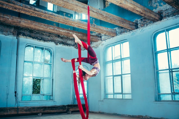 Agraciado gimnasta realizando ejercicio aéreo con telas rojas en azul antiguo loft