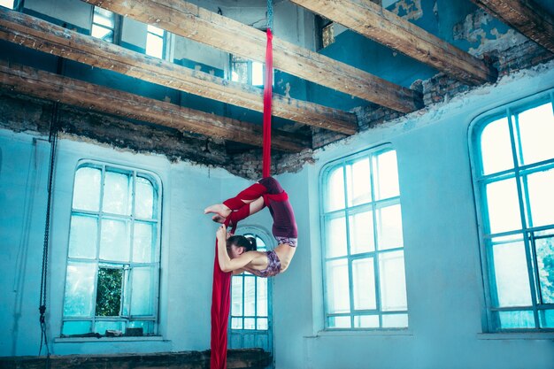 Agraciado gimnasta realizando ejercicio aéreo con telas rojas en azul antiguo loft