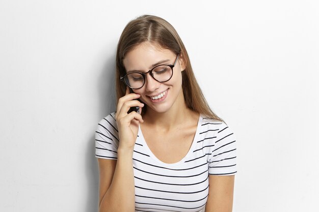 Agradable adolescente mujer tímida con largo cabello suelto mirando hacia abajo y sonriendo ampliamente mientras habla por teléfono móvil