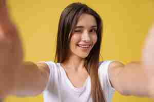 Foto gratuita agradable atractiva chica haciendo selfie en estudio y riendo. mujer joven guapa con cabello castaño tomando una foto de sí misma sobre fondo amarillo brillante.