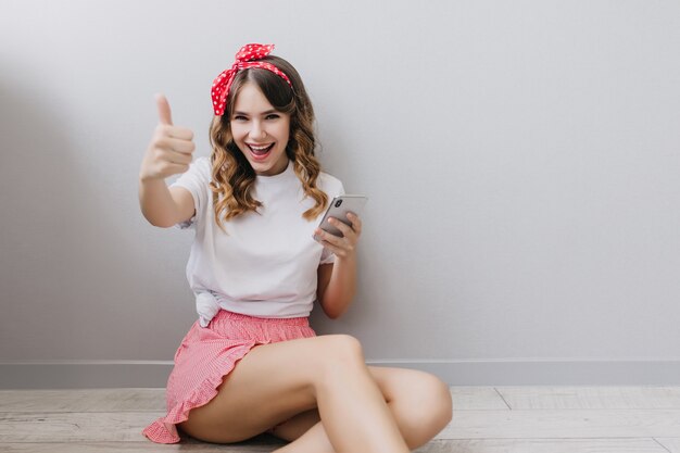 Agradable chica caucásica con cinta roja en cabello oscuro sentada en el suelo en casa. Filmación en interiores de modelo femenino sonriente en pantalones cortos rosas.